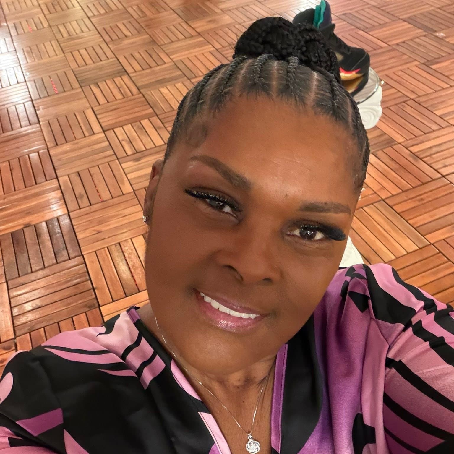 Smiling woman with braided hair in a bun wearing a purple and black patterned top.