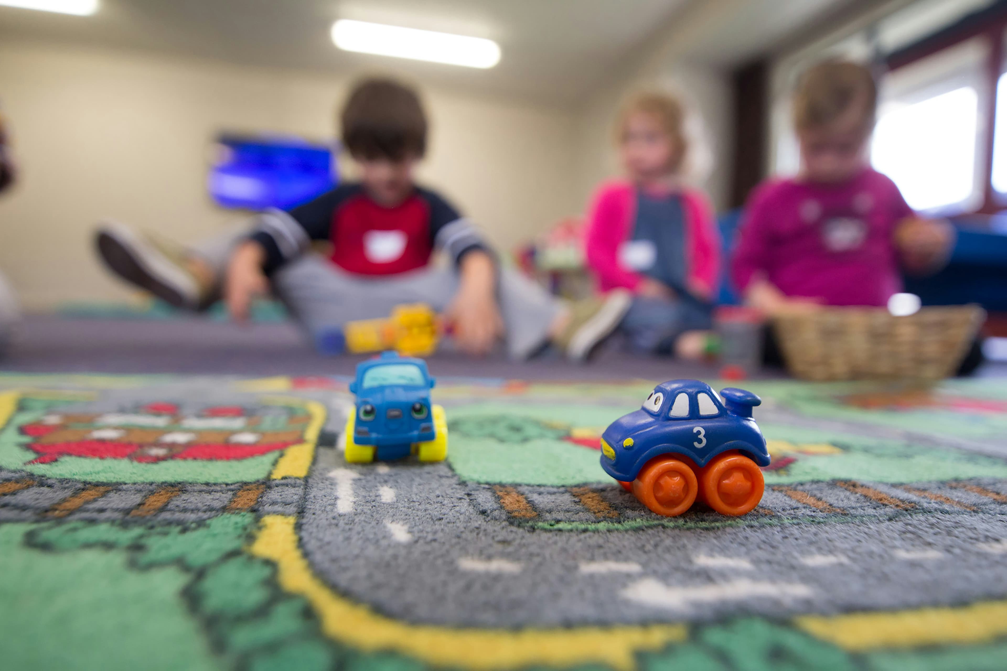 Two toy cars on a road-patterned rug with blurred children playing in the background.