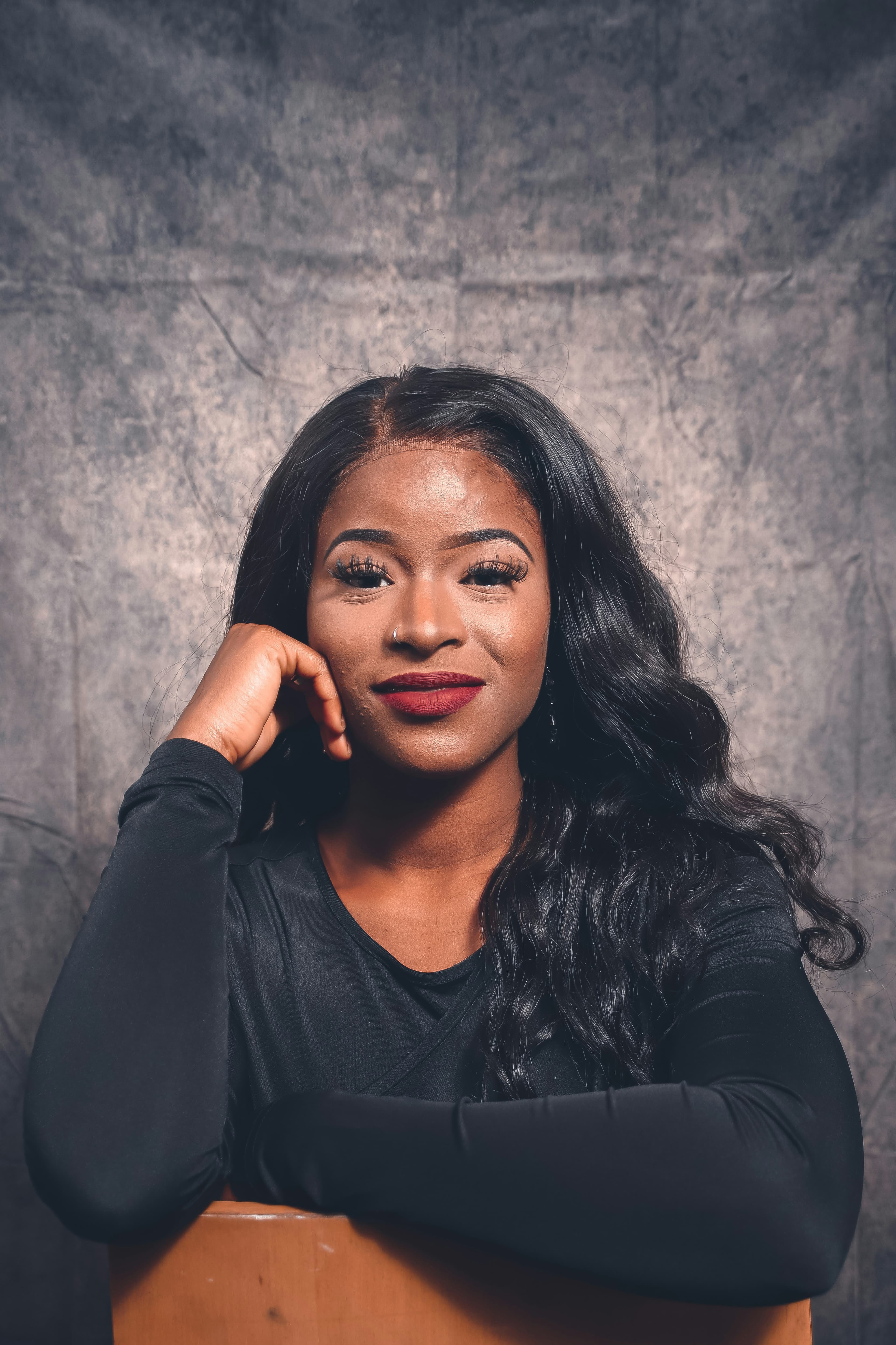 Black woman with long wavy hair and red lipstick posing against a grey textured background.