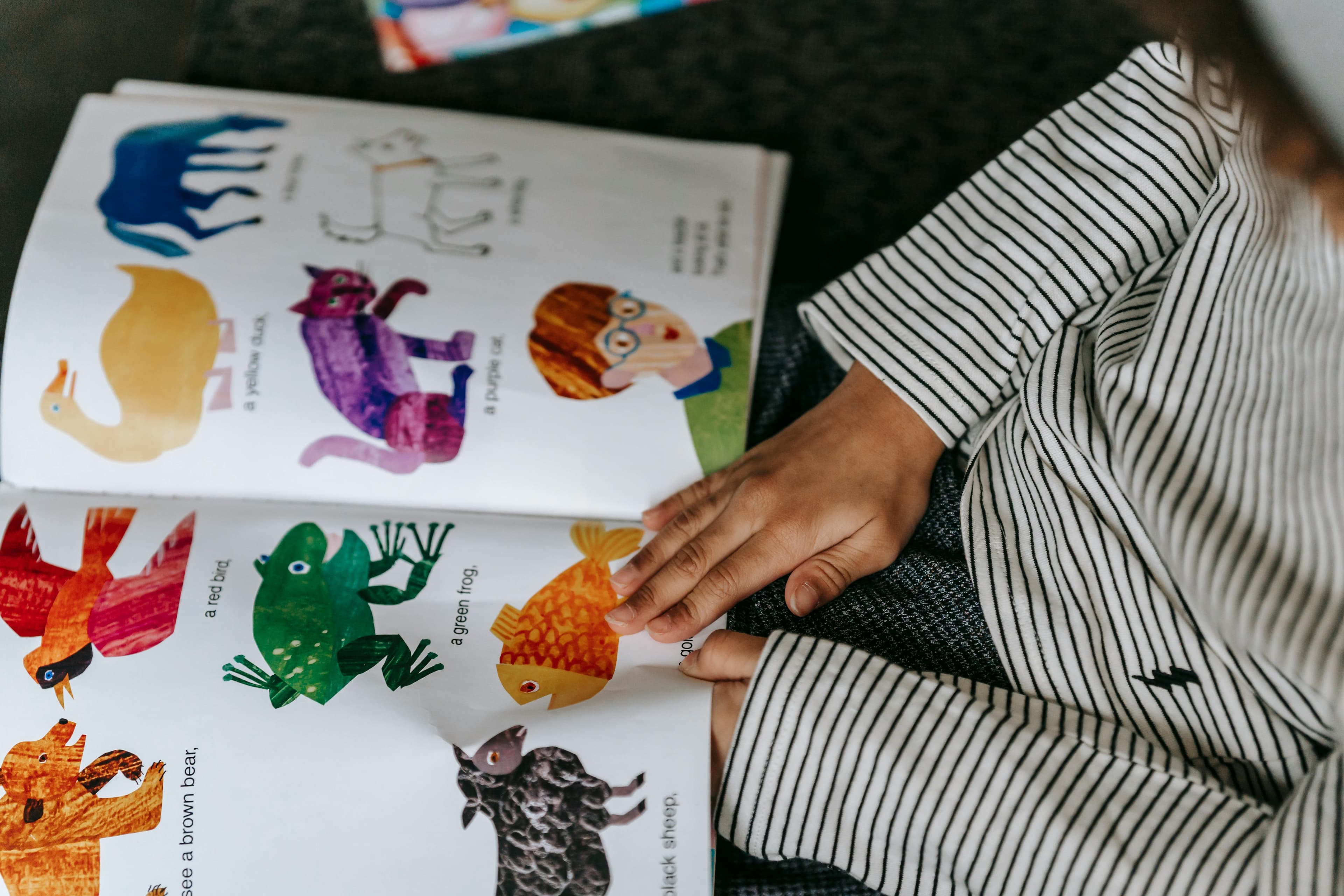 Child in a striped shirt points at colorful animal illustrations in an open picture book.